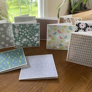 May include: A collection of folded greeting cards with various patterned designs. The cards feature floral, bird, and geometric patterns in colors like green, blue, gray, and brown. The cards are displayed on a wooden surface.