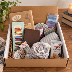 May include: A cardboard gift box containing a brown paper-wrapped book labeled Blind Date with a Book, a colorful stack of books bookmark, a dark brown notebook, a gray ribbed mug, fuzzy gray socks, Milano dark chocolate cookies, and stickers.