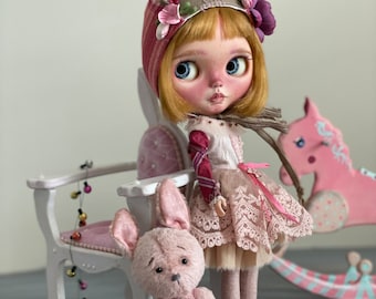 May include: A doll with blonde hair wearing a pink and red hat with floral accents, a pink and white dress, and brown shoes. The doll is holding a pink bunny plush toy. The doll is sitting on a white chair with a pink cushion. There are pink and white decorations on the chair.
