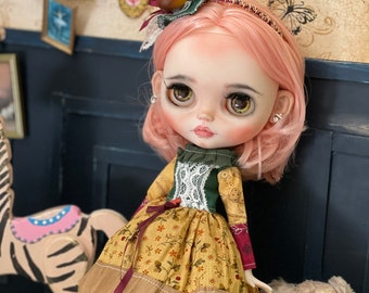 May include: A doll with pink hair wearing a yellow and brown dress with a green bodice and white lace. The doll is wearing a headband with an acorn on top. The doll is standing in front of a blue wall with a wooden rocking horse and a teddy bear.