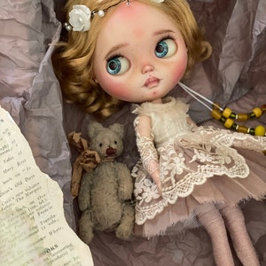 May include: A vintage-style doll with blonde hair, blue eyes, and a floral headband. The doll wears a cream-colored lace dress and sits with a small gray teddy bear. The doll is in a box with torn paper and text from a book.