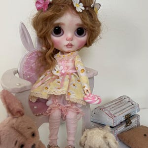May include: A doll with long curly brown hair wearing a yellow and white floral dress, a pink headband with butterflies and flowers, and pink shoes. The doll is sitting on a white chair with a pink cushion. There are two teddy bears, one pink and one white, sitting on the floor in front of the doll.