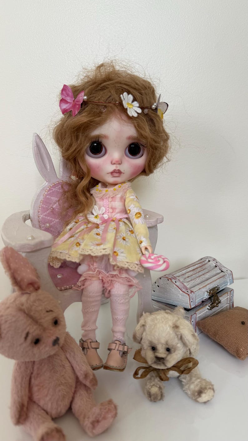 May include: A doll with long curly brown hair wearing a yellow and white floral dress, a pink headband with butterflies and flowers, and pink shoes. The doll is sitting on a white chair with a pink cushion. There are two teddy bears, one pink and one white, sitting on the floor in front of the doll.