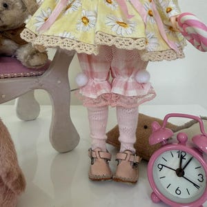 May include: A doll wearing a yellow and pink gingham dress with a daisy print, pink tights, and pink shoes. The doll is holding a candy cane and sitting on a small chair.