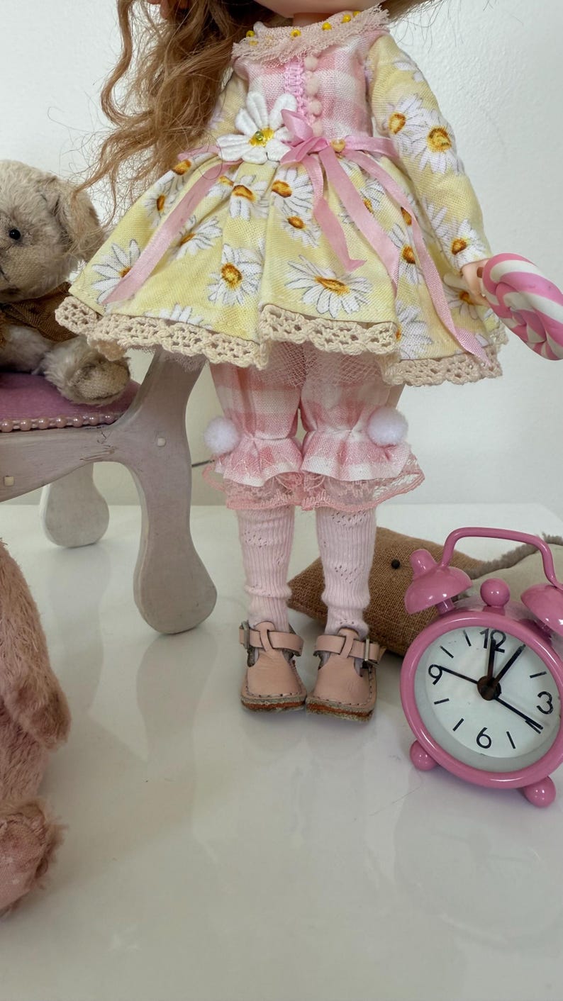 May include: A doll wearing a yellow and pink gingham dress with a daisy print, pink tights, and pink shoes. The doll is holding a candy cane and sitting on a small chair.