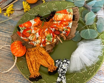 May include: A green ceramic plate featuring a fall-themed doll outfit. The outfit consists of a pumpkin-print dress, brown trousers, and a white tulle skirt. A small pumpkin is also on the plate.