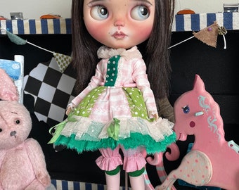 Può includere: Una bambola con capelli scuri e occhi blu, indossa un vestito rosa e verde con finiture in pizzo, leggings rosa e scarpe a forma di rana. La bambola è in piedi su una base a forma di fiore sopra una scatola di legno con un disegno di rose.