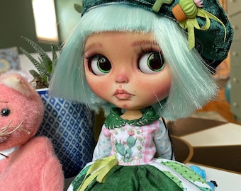 May include: A green and white doll with blue hair wearing a green beret with a cactus pin. The doll is wearing a green and white dress with a cactus print. The doll is sitting on a white surface.