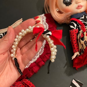 May include: A white pearl headband with a red and black bow. The headband is made of a thin wire and has a black elastic band.