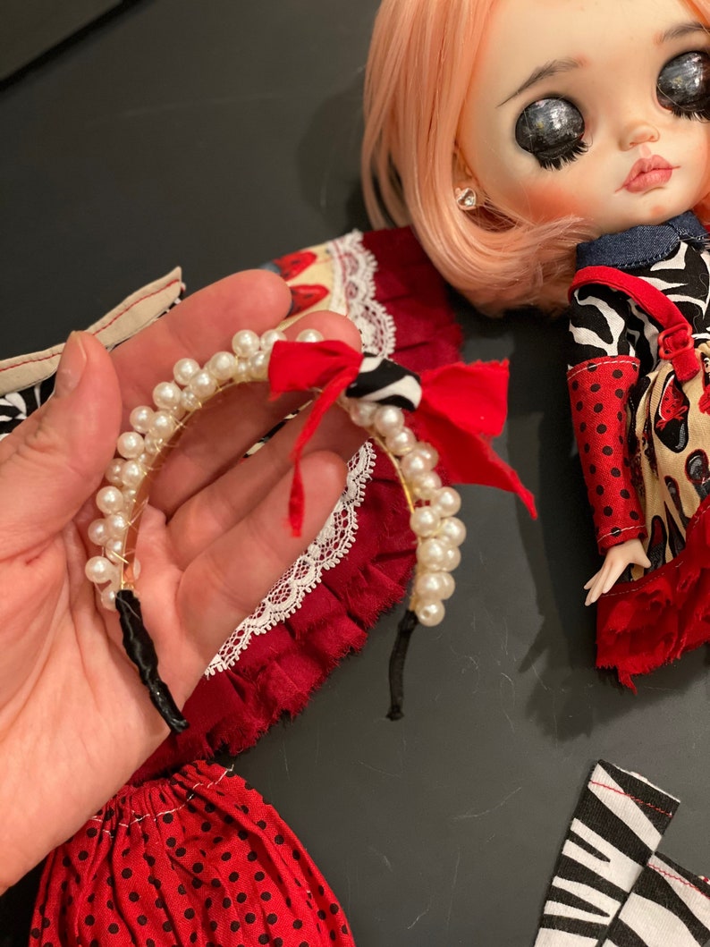 May include: A white pearl headband with a red and black bow. The headband is made of a thin wire and has a black elastic band.