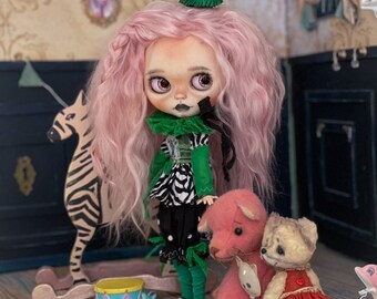 May include: A pink haired doll wearing a green and black clown costume with a green hat. The doll is holding a pink teddy bear and there is a brown teddy bear with a red dress sitting next to it. There is a zebra rocking horse and a small drum in the background.