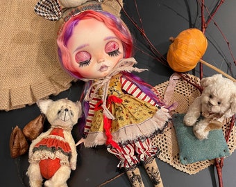 May include: A doll with pink and purple hair wearing a red and white striped dress and a brown and white plaid hat. The doll is lying on a black surface with a white teddy bear and a brown pumpkin.