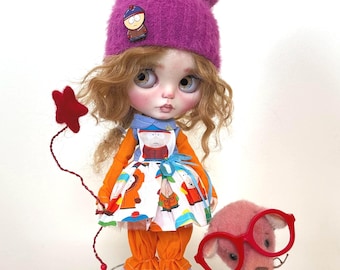 Può includere: Una bambola con capelli biondi ricci che indossa un cappello di pelliccia viola con un pon pon rosso, un vestito bianco con una stampa di cartoni animati, pantaloni arancioni e calze a righe blu e bianche. La bambola tiene una stella rossa su un bastone. Un orsacchiotto di peluche rosa con occhiali rossi è seduto accanto alla bambola. Un piccolo contenitore di popcorn è in primo piano.