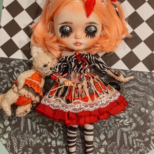 May include: A doll with pink hair, wearing a red and white dress with a black and white zebra print top, holding a wand. The doll is wearing black and white striped stockings and red shoes. The doll is sitting on a black and white checkered background with a teddy bear.