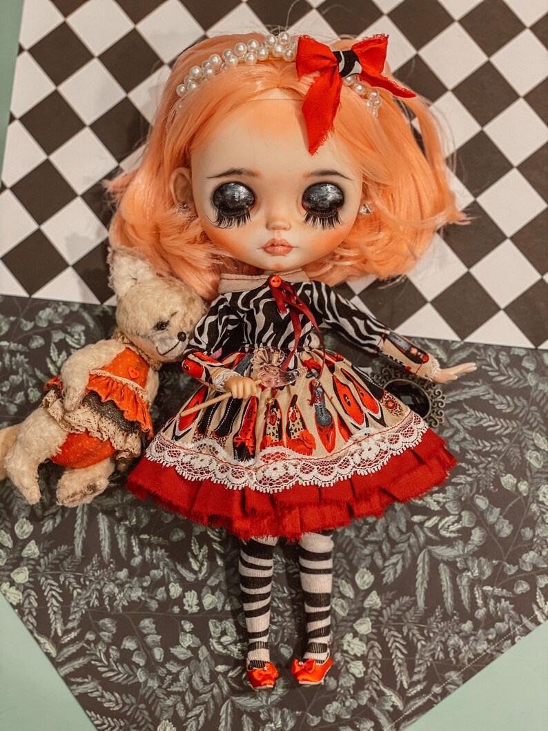 May include: A doll with pink hair, wearing a red and white dress with a black and white zebra print top, holding a wand. The doll is wearing black and white striped stockings and red shoes. The doll is sitting on a black and white checkered background with a teddy bear.