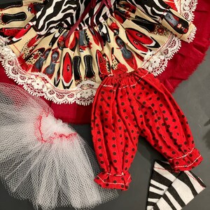 May include: A doll outfit set with a red and black zebra print dress, red polka dot pants, and black and white striped stockings. The dress has a white lace trim and a red bow. The outfit is perfect for a doll with a 14-inch body.