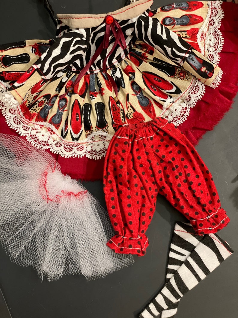 May include: A doll outfit set with a red and black zebra print dress, red polka dot pants, and black and white striped stockings. The dress has a white lace trim and a red bow. The outfit is perfect for a doll with a 14-inch body.
