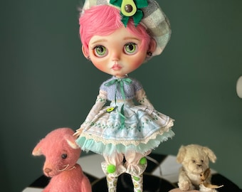 May include: A pink doll with green eyes and pink hair wearing a plaid beret, a blue and white dress, and avocado-themed socks. The doll is standing on a black and white checkered surface with two plush toys, a pink cat and a beige dog.