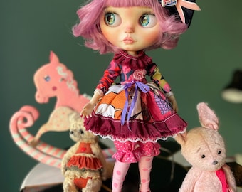 Può includere: Una bambola rosa con occhi azzurri e capelli rosa che indossa un vestito rosso e nero, un berretto blu con un bottone rosa e blu e calze rosa. La bambola è in piedi su un tavolo bianco e nero con due animali di peluche, un coniglietto rosa e un orsacchiotto marrone.