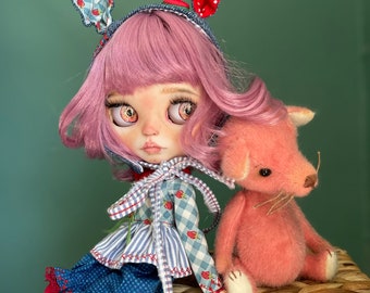 Può includere: Una bambola con i capelli rosa che indossa un vestito a quadri blu e bianchi con un cerchietto con orecchie di coniglio a pois rossi e bianchi. La bambola è seduta su un cesto intrecciato con un giocattolo di peluche rosa accanto a lei.