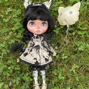 May include: A black-haired doll with large blue eyes is wearing a black and white cat-themed dress and a black cat ear headband. The doll is holding a white cat-shaped balloon.
