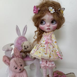 May include: A doll with long brown hair wearing a yellow and white floral dress with a pink ribbon. The doll is sitting on a white chair with bunny ears. There are two pink teddy bears and a pink alarm clock on the table.