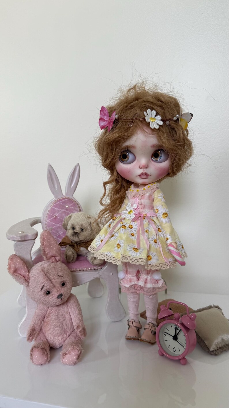 May include: A doll with long brown hair wearing a yellow and white floral dress with a pink ribbon. The doll is sitting on a white chair with bunny ears. There are two pink teddy bears and a pink alarm clock on the table.