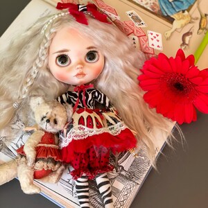 May include: A white haired doll with green eyes wearing a red and black dress and white stockings. The doll is sitting on an open book with playing cards scattered around it. A red flower is in the background.