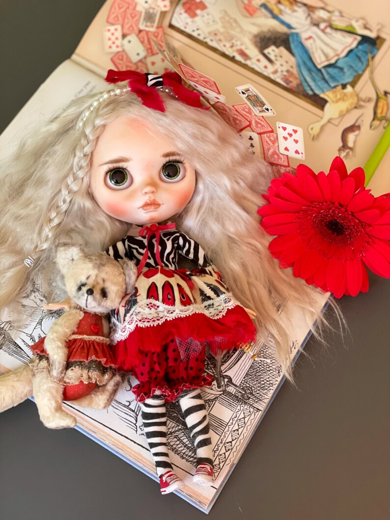 May include: A white haired doll with green eyes wearing a red and black dress and white stockings. The doll is sitting on an open book with playing cards scattered around it. A red flower is in the background.