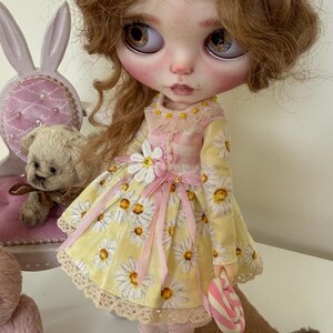 May include: A doll with long blonde curly hair wearing a yellow daisy print dress with a pink ribbon and a white daisy on the bodice. The doll is holding a pink and white candy cane and is wearing brown shoes. The doll is sitting on a brown surface with a pink alarm clock and a pink teddy bear.