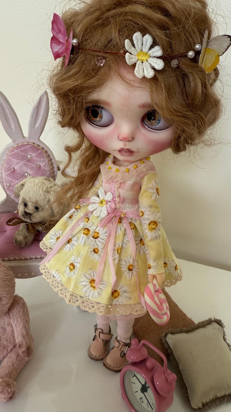 May include: A doll with long blonde curly hair wearing a yellow daisy print dress with a pink ribbon and a white daisy on the bodice. The doll is holding a pink and white candy cane and is wearing brown shoes. The doll is sitting on a brown surface with a pink alarm clock and a pink teddy bear.
