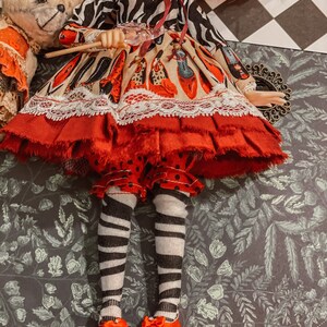 May include: A doll wearing a red and white dress with a black and white zebra print top, black and white striped tights, and red shoes. The doll is holding a wand and is standing on a green and brown patterned surface.