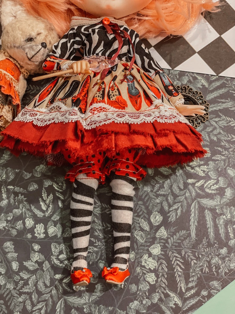 May include: A doll wearing a red and white dress with a black and white zebra print top, black and white striped tights, and red shoes. The doll is holding a wand and is standing on a green and brown patterned surface.