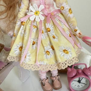 May include: A yellow and white daisy print dress with pink ribbon accents and a white daisy flower embellishment. The dress has a lace trim and the doll is wearing pink socks and brown shoes.