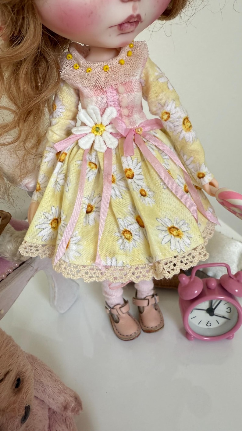 May include: A yellow and white daisy print dress with pink ribbon accents and a white daisy flower embellishment. The dress has a lace trim and the doll is wearing pink socks and brown shoes.