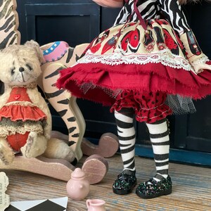 May include: A doll wearing a red and white dress with a zebra print top, black and white striped tights, and black shoes with glitter. The doll is sitting on a rocking horse with a teddy bear sitting on the horse's back.