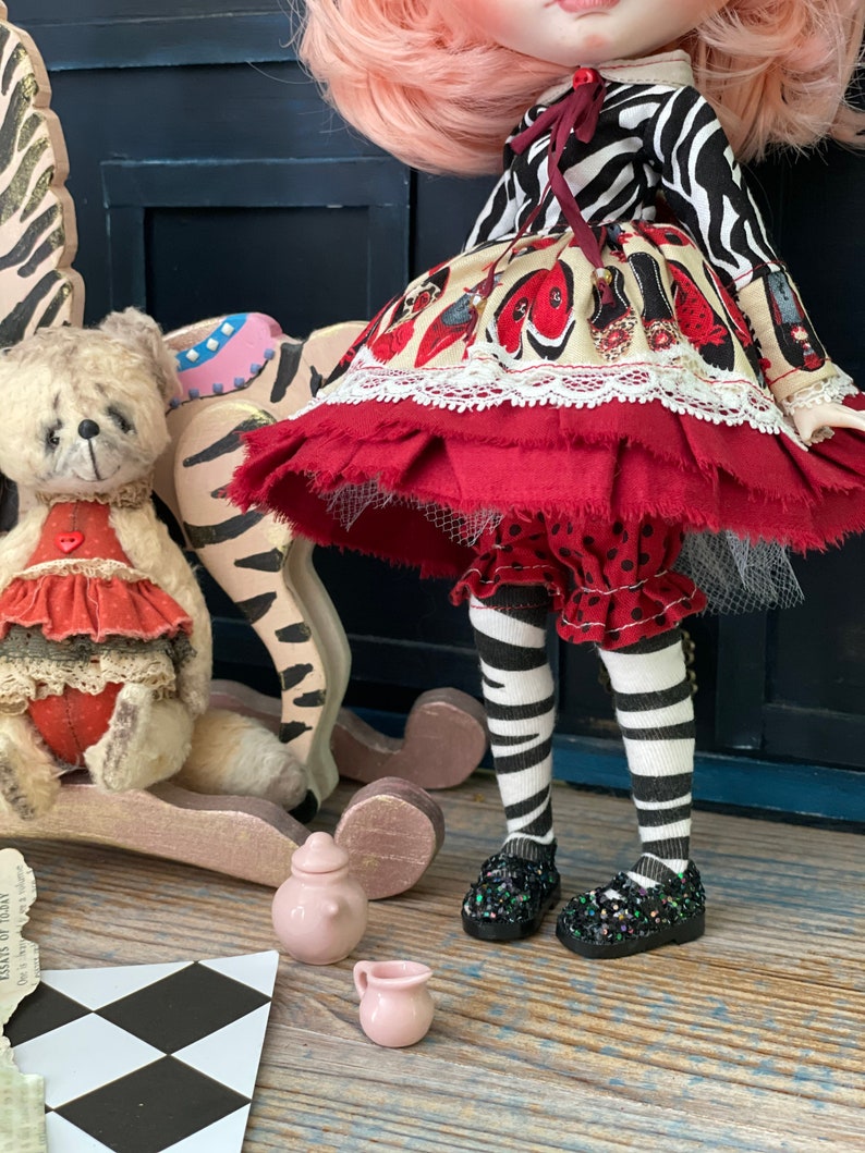 May include: A doll wearing a red and white dress with a zebra print top, black and white striped tights, and black shoes with glitter. The doll is sitting on a rocking horse with a teddy bear sitting on the horse's back.
