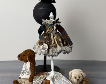 May include: A steampunk-themed display featuring a miniature dress on a mannequin, a top hat with goggles, and two plush dogs. The dress is adorned with gears and lace. The dogs wear accessories. A pair of striped trousers and socks are also present.