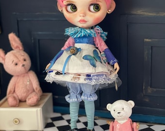 May include: A pink haired doll wearing a blue and white dress with a butterfly design. The doll is holding a small stuffed animal and is standing on a black and white checkered floor.