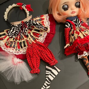May include: A doll wearing a red and black dress with a white and black zebra print. The doll has pink hair and is wearing a headband with pearls. The doll is also wearing red and black polka dot pants and black and white striped stockings.