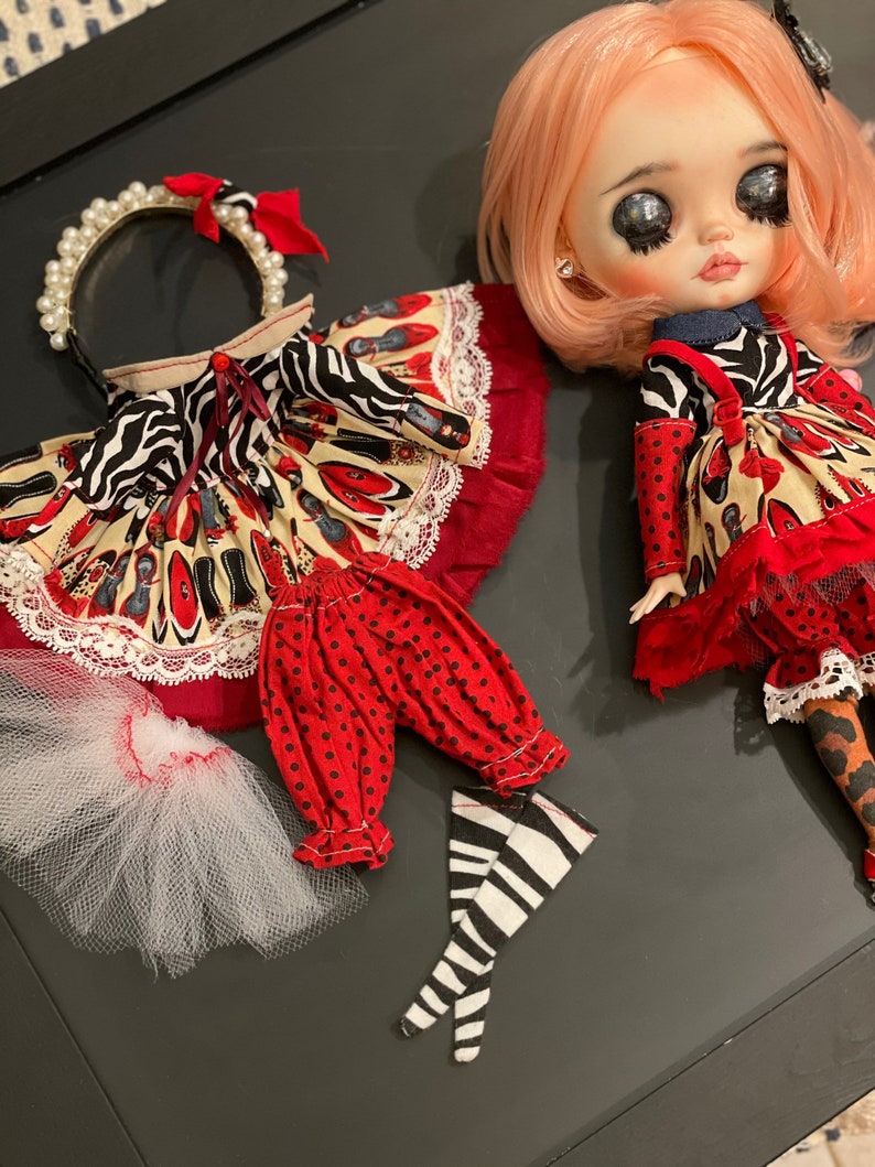 May include: A doll wearing a red and black dress with a white and black zebra print. The doll has pink hair and is wearing a headband with pearls. The doll is also wearing red and black polka dot pants and black and white striped stockings.