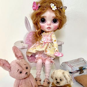 May include: A doll with long, curly red hair wearing a yellow and white floral dress and pink tights sits on a white bunny-shaped chair. The doll is holding a pink lollipop. There are two stuffed animals, a pink bunny and a cream-colored dog, sitting on the floor in front of the doll.