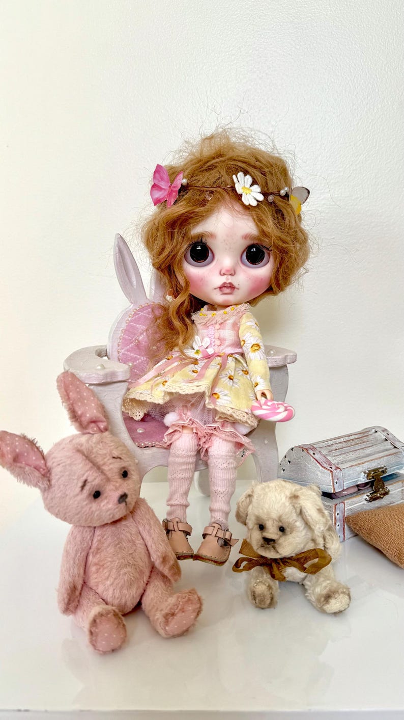 May include: A doll with long, curly red hair wearing a yellow and white floral dress and pink tights sits on a white bunny-shaped chair. The doll is holding a pink lollipop. There are two stuffed animals, a pink bunny and a cream-colored dog, sitting on the floor in front of the doll.