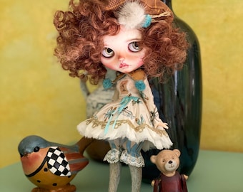 Può includere: Una bambola dettagliata con capelli rossi ricci, che indossa un vestito fantasia, un cappello e stivali. Un uccello di ceramica e un piccolo orsacchiotto sono posti vicino alla bambola. Un vaso scuro con fiori gialli è sullo sfondo.
