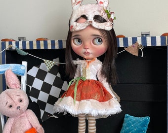 Può includere: Una bambola con una maschera di coniglio bianca e un vestito multicolore sta in piedi su una scatola di legno con la parola "sprout" scritta su di essa. La bambola è circondata da animali di peluche, tra cui un coniglio rosa che tiene una carota e un coniglio bianco che indossa un vestito rosso.