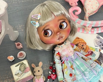 May include: A doll with brown skin and blonde hair wearing a pastel blue and pink dress with a white lace trim. The doll is sitting on a wooden surface with a pink rocking horse, a small wooden box, and a teacup and saucer.