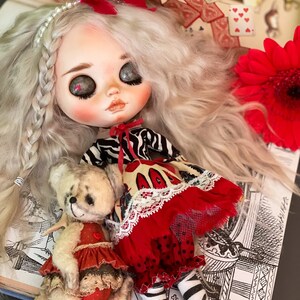 May include: A doll with white hair and a red and black striped dress, holding a teddy bear wearing a red and white dress. The doll is sitting on a book with a black and white illustration.