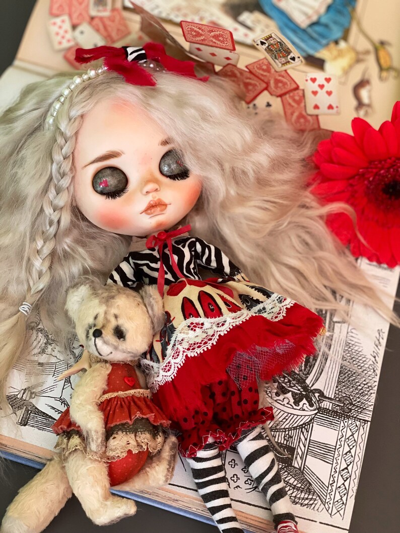 May include: A doll with white hair and a red and black striped dress, holding a teddy bear wearing a red and white dress. The doll is sitting on a book with a black and white illustration.