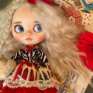 May include: A doll with long blonde hair wearing a red and white dress with a black and white zebra print top. The doll is wearing a white lace collar and a red bow with a white and black striped ribbon. The doll is holding a white furry object in her left hand. The doll is sitting on a table with a background of playing cards and a floral pattern.