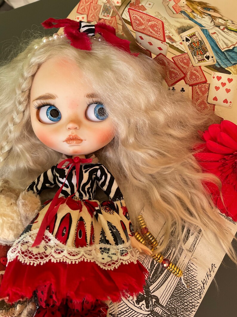 May include: A doll with long blonde hair wearing a red and white dress with a black and white zebra print top. The doll is wearing a white lace collar and a red bow with a white and black striped ribbon. The doll is holding a white furry object in her left hand. The doll is sitting on a table with a background of playing cards and a floral pattern.